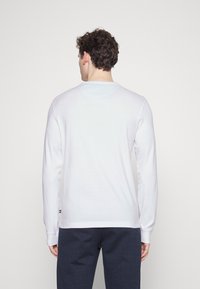 Felpa bianca a maniche lunghe con colletto rotondo e polsini a costine, caratterizzata da una texture liscia e un logo accentato sull'orlo.