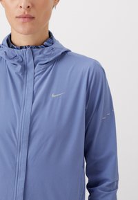 Kevyt violetti Nike-takki, jossa on vetoketjullinen etuosa, huppu ja heijastava logo. Takissa on sileä pinta ja hienovaraiset yksityiskohdat hihoissa.