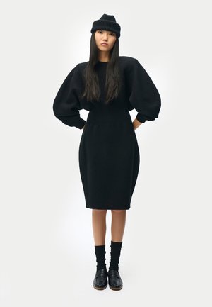 Femme debout, mains sur les hanches, portant une robe en maille noire avec des manches bouffantes, un bonnet noir, des chaussettes noires et des mocassins noirs.