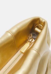 Wnętrze złotej skórzanej torby, charakteryzujące się gładką fakturą, widocznym zamkiem błyskawicznym i metalową metką z logo. Strukturalny kształt z miękkim otwarciem.