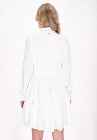 Robe blanche à manches longues, avec un col haut, une taille ceinturée et une jupe à volants. Tissu lisse avec une texture subtile ; fermée à l'arrière.