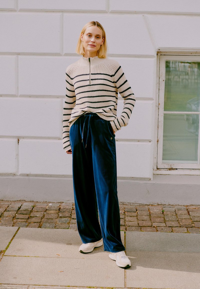 Prúžkovaný sveter s vysokým golierom v krémovej a čiernej farbe; v kombinácii s námorníckymi zamatovými nohavicami s širokými nohavicami. Biele tenisky dotvárajú outfit.