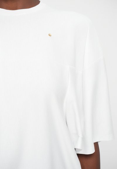 Persoon in een eenvoudig wit T-shirt met een kleine gouden speld op de bovenborst, zichtbaar van de schouders tot halverwege de torso.