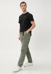 Zwarte T-shirt, olijfgroene cargobroek met zakken en een verstelbare tailleband, gecombineerd met witte sneakers. Model in een staande pose.