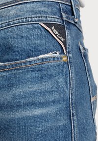 Vaqueros de mezclilla azul con textura desgastada, que presentan una etiqueta negra con texto blanco y costura roja. Detalle de remache dorado en el bolsillo.