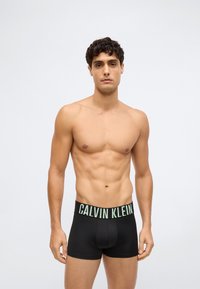 Black Calvin Klein boxer briefs with a green logo on the waistband, smooth texture, and fitted design, modeled by a shirtless man.