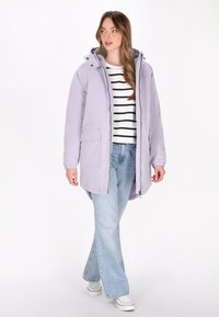 Gai lavandas parka ar rāvējslēdzēju priekšpusē, regulējamu kapuci, elastīgām piedurknēm un divām priekšējām kabatām, uzvilkta virs svītrainas krekla un denim džinsiem.