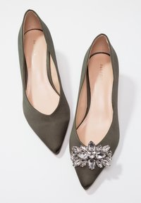 Paire de ballerines vert olive à bout pointu, dont une ornée d'un grand ensemble décoratif de pierres translucides semblables à des cristaux.