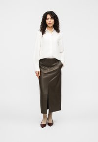 Λευκό πουκάμισο με κουμπιά και μακριά μανίκια συνδυασμένο με μια σκουροπράσινη δερμάτινη φούστα με πλαϊνή σχισμή, μπεζ κάλτσες και σκουρόκόκκινα μυτερά παπούτσια.