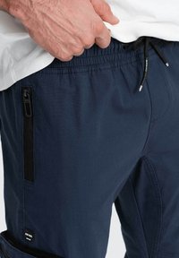 Pantalons bleu marine en tissu léger, dotés d'une taille élastique, d'une poche zippée avec des finitions noires et d'une étiquette de marque visible.