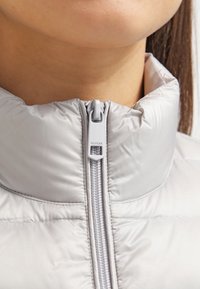 Jasnoszara, ocieplana kurtka z wysokim kołnierzem i zamkiem błyskawicznym z przodu. Posiada gładką, błyszczącą tkaninę oraz suwak oznaczony napisem "HILFIGER."