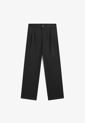 Personne portant un pantalon noir à jambes larges, une ceinture noire, un haut noir et des chaussures noires, debout de profil sur un fond clair uni.