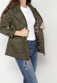 Kobieta w oliwkowej kurtce z kratkowanym wnętrzem, białej bluzce i niebieskich dżinsach z haftem w kwiaty, trzymająca kurtkę otwartą z przodu.