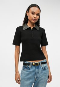 Melns ribots īsajiem piedurknēm topiņš ar rāvējslēdzēju priekšā un studētu apkakli. Valkāts ar gaiši ziliem džinsiem un melnu jostu.