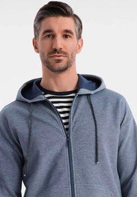 Haut à capuche zippé gris avec un intérieur contrastant bleu marine, doté d'une capuche avec cordon de serrage et d'une poche kangourou, porté sur une chemise rayée noir et blanc.