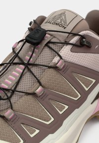 Salomon atletisk sko med en lys brun mesh-overdel, pink accenter og et hurtigt snøresystem. Bemærkelsesværdigt skråt design på siderne.