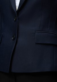 Κοντινή λήψη ενός σκουρόχρωμου μπλε σακακιού με δύο μαύρα κουμπιά και τσέπη με καπάκι, που φοριέται πάνω από ένα λευκό πουκάμισο.