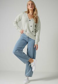 Cardigan lavorato a maglia grigio chiaro con scollatura a V, cinque bottoni neri e polsini a costine indossato sopra jeans a gamba ampia blu e sneakers bianche.