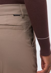 Κοντινό πλάνο στην τσέπη και τη θηλιά ζώνης ενός καφέ παντελονιού με ετικέτα Peak Performance, μαζί με ένα χέρι και μανίκι από ένα ριμπ πλεκτό σκούρο καφέ πουλόβερ.