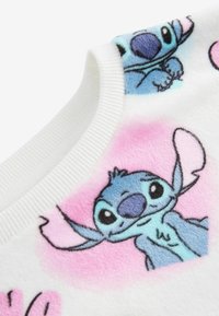 Valkoinen pörröinen neulepaita, jossa on värikkäitä Stitch-hahmon kuvioita vaaleanpunaisella taustalla. Ribattu kaula-aukko ja pehmeä rakenne.