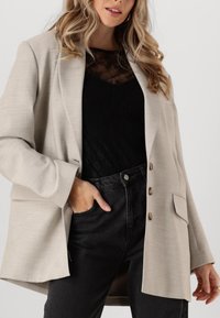 Blazer beige con un corte relajado, textura suave y cuatro acentos de botones dorados, combinado con una blusa de encaje negra y jeans de tiro alto negros.