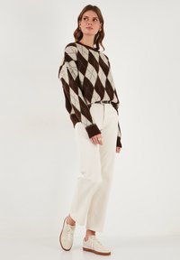 Rjav in krem argyle pleten pulover, sproščenega kroja, v kombinaciji z belimi hlačami ravnega kroja in svetlobrnimi športnimi čevlji. Teksturni, sproščen stil.