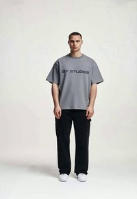 Jovem está de pé, de frente, vestindo uma t-shirt oversized cinza com o texto "2Y STUDIOS", calças pretas e ténis brancos, num fundo simples.