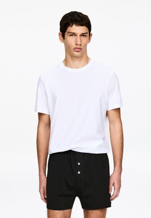 Jeune homme aux cheveux courts et foncés portant un t-shirt blanc uni et un boxer noir boutonné, debout devant un fond blanc.