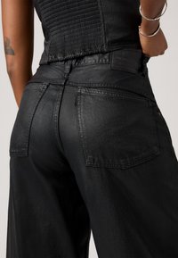 Personne portant un jean noir taille haute et un haut texturé noir, montrant les poches arrière et un tatouage sur son bras.
