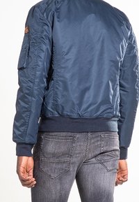 Personne portant une veste bomber bleu marine et un jean gris en denim, vue de dos, les mains détendues le long du corps.