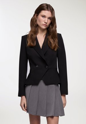 Blazer nero su misura con rever a punta, doppi bottoni e vita peplum strutturata. Abbinato a una gonna grigia plissettata. Tessuto dalla consistenza liscia.