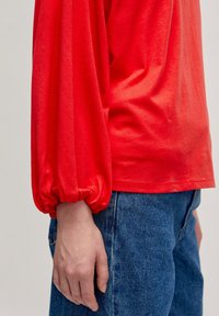Rotes langärmliges Oberteil mit gerafften Manschetten, aus weichem Stoff gefertigt. Kombiniert mit blauen Jeans im klassischen Schnitt mit sichtbaren Nähdetails.