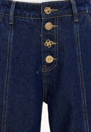 Hochgeschnittene blaue Jeanshose mit goldenen Dekoknöpfen, Kontrastnähten und einem leicht strukturierten Stoff.