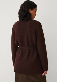 Hnědý cardigan s dlouhým rukávem vyrobený z měkkého, texturovaného materiálu, se zavazovacím páskem v pase a délkou do boků. Bez viditelných zapínání.