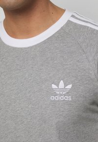 Close-up van een persoon die een grijze Adidas t-shirt draagt met witte drie strepen op de schouder en het Adidas-logo op de borst.