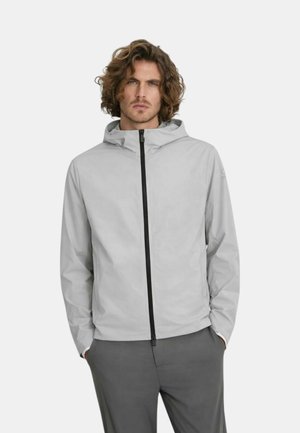 Uomo con capelli ricci che indossa una giacca con cappuccio grigio chiaro con cerniera nera, mani nelle tasche, in piedi davanti a uno sfondo uniforme.