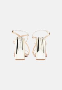 Πέδιλα με ψηλό τακούνι σε στυλ με λουράκια, με μεταλλικό χρυσό τακούνι σε σχήμα μπλοκ, διαφανείς πέτρες στα λουράκια και βάση σε γυμνό χρώμα.