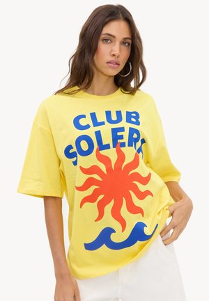Jeune femme aux longs cheveux bruns porte un T-shirt jaune oversize avec un soleil rouge, des vagues bleues et le texte « CLUB SOLERO », associé à un pantalon blanc.