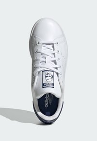 White leather sneaker with a rounded toe, lace-up front, and navy accents. Features a graphic label on the tongue and perforated sides.