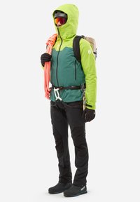 Veste de plein air verte et noire avec capuche, pantalon renforcé et équipement d'escalade. Le modèle tient une corde orange, portant des gants et des lunettes de protection.