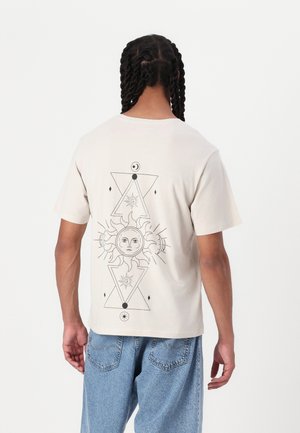 Osoba z warkoczami, ubrana w beżowy t-shirt z czarnym słońcem i geometrycznymi wzorami na plecach oraz niebieskie dżinsy.