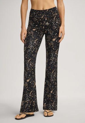 Femme portant des leggings évasés taille haute à imprimé abstrait noir et beige, avec des tongs noires, debout devant un fond neutre.