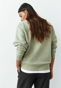 Gaiši zaļa sporta džemperis ar nolaižamām plecu līnijām, ribveida aprocēm un apakšmalu. Slāņots pār baltu topu, kombinēts ar tumšām svītrainām biksēm.