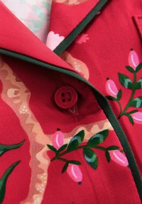 Cămașă din țesătură roșie cu modele florale și accente verzi, guler decupat și detaliu cu nasture roșu. Textură netedă, design jucăuș.