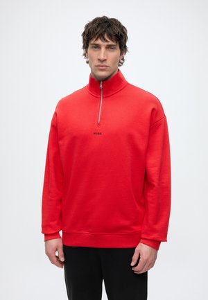 Mann mit dunklen Haaren, der einen roten Zip-Pullover mit „HUGO“-Logo trägt und vor einem schlichten weißen Hintergrund steht.