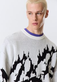 Pull en maille blanc avec un motif abstrait noir audacieux. Présente un col rond et une couche violette en dessous. Texture douce.