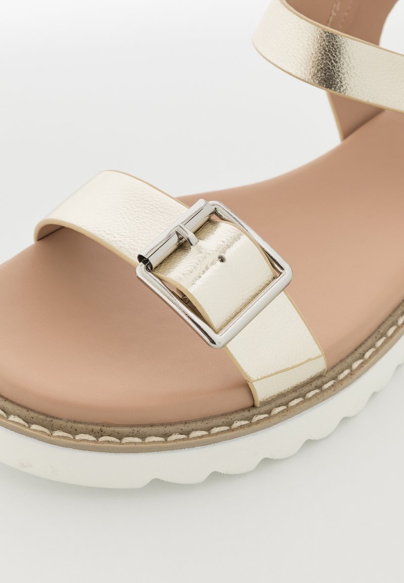 Sandale métallique dorée avec une grande boucle argentée, une large sangle et une semelle intérieure texturée. Semelle en caoutchouc blanche avec un léger détail de couture.