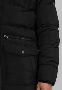 Černá péřová bunda s kapsou na zip a klopovou kapsou, charakterizovaná hladkou texturou, prošívanými liniemi a elastickými manžetami.