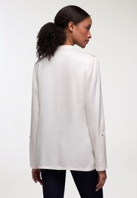 Blusa di seta bianca con maniche lunghe, caratterizzata da una texture liscia, polsini abbottonati e una vestibilità rilassata, mostrata da un'angolazione posteriore.