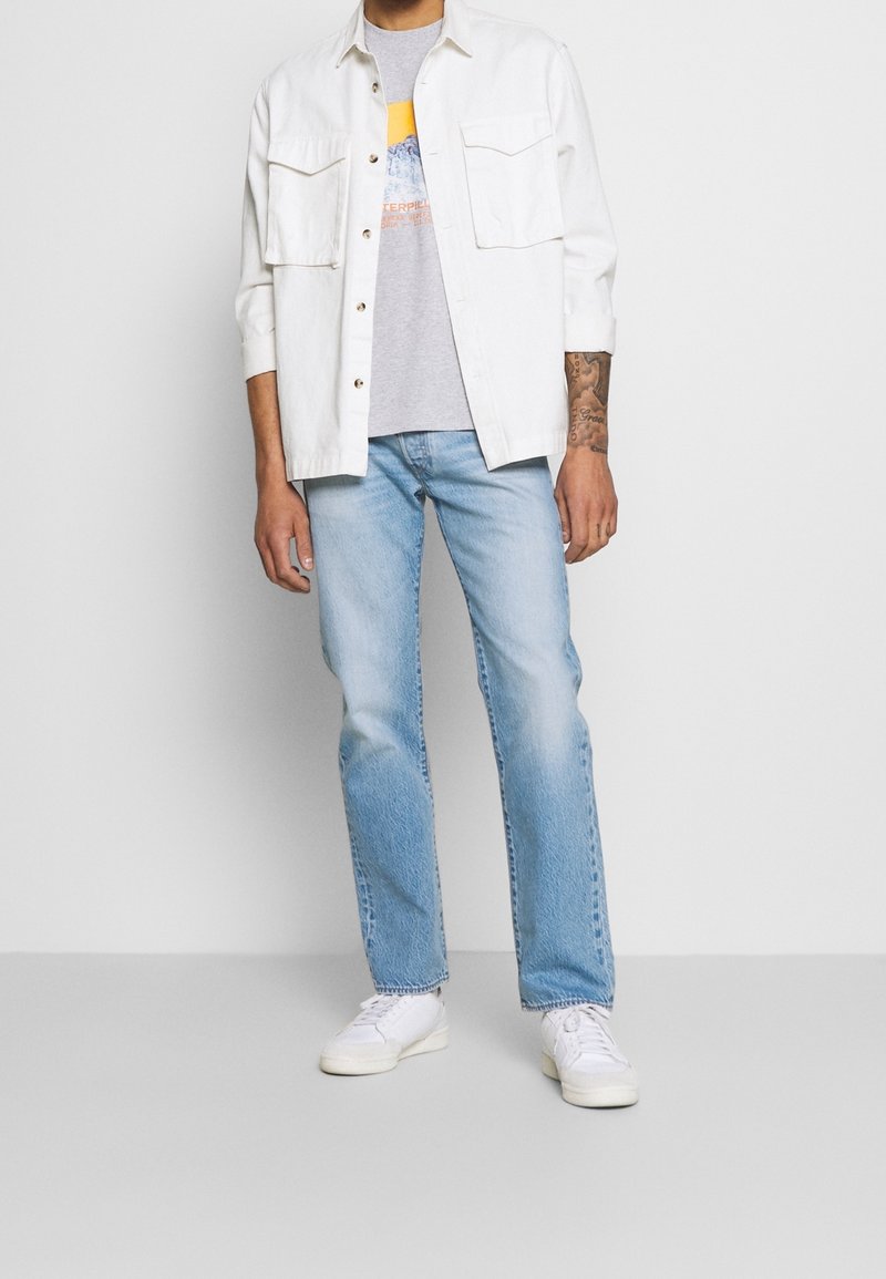 Vit skjorta med krage över en grå grafisk t-shirt, i kombination med ljusblå jeans och vita sneakers. Skjortan har fickor.